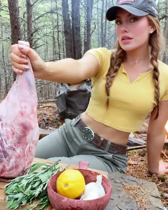 A raw leg of lamb held up over a wooden prep board with fresh herbs, a lemon and garlic, deep in a pine forest